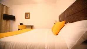 A hotel room with a large bed, a flat-screen TV mounted on the wall, a small desk with a chair, and a framed picture on the wall. - Hotel Illiyin (Dakar)