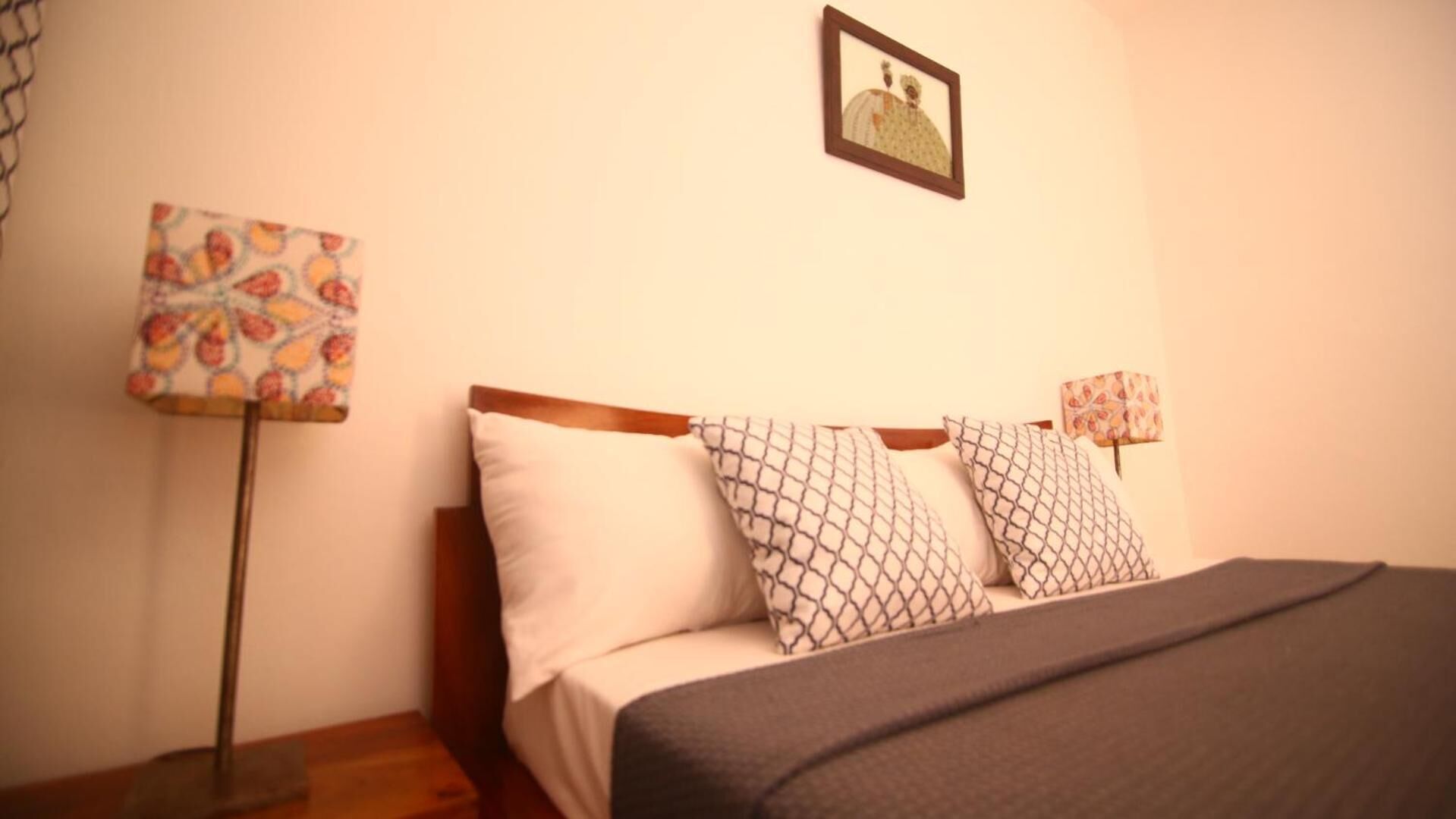 Una cama bien hecha con almohadas estampadas, una mesita de noche con lámpara y un cuadro en la pared.
