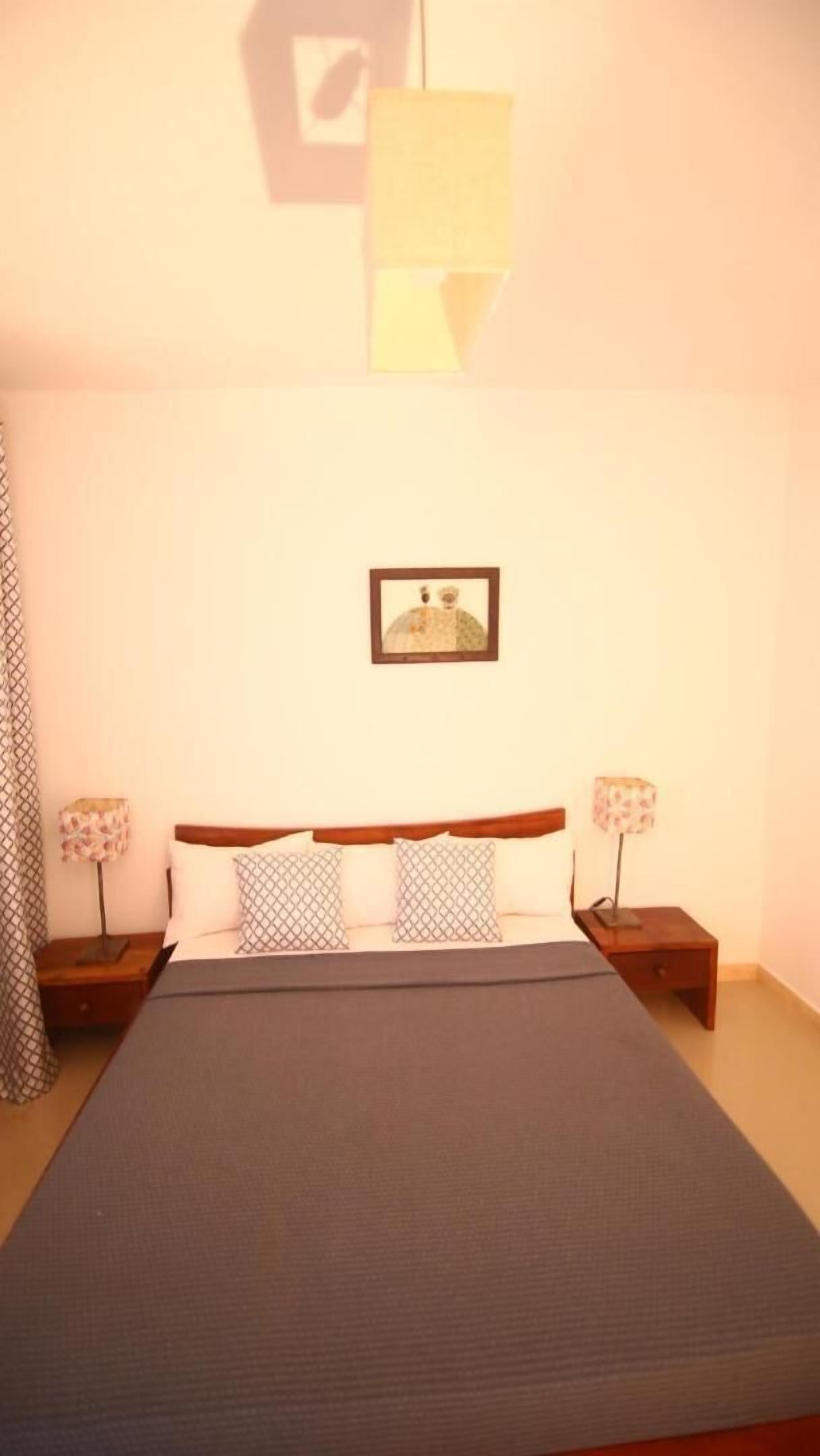 Un dormitorio con una cama, dos mesitas de noche, una lámpara en cada mesita y un cuadro en la pared, además de una lámpara de techo con diseño geométrico.