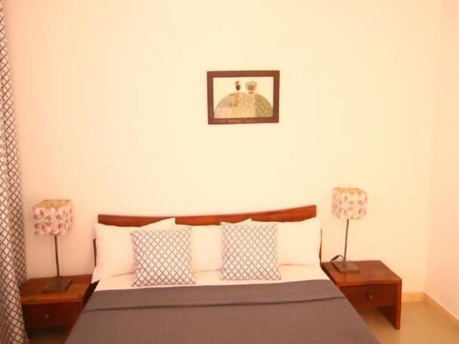 Un dormitorio con una cama, dos mesitas de noche, una lámpara en cada mesita y un cuadro en la pared, además de una lámpara de techo con diseño geométrico.