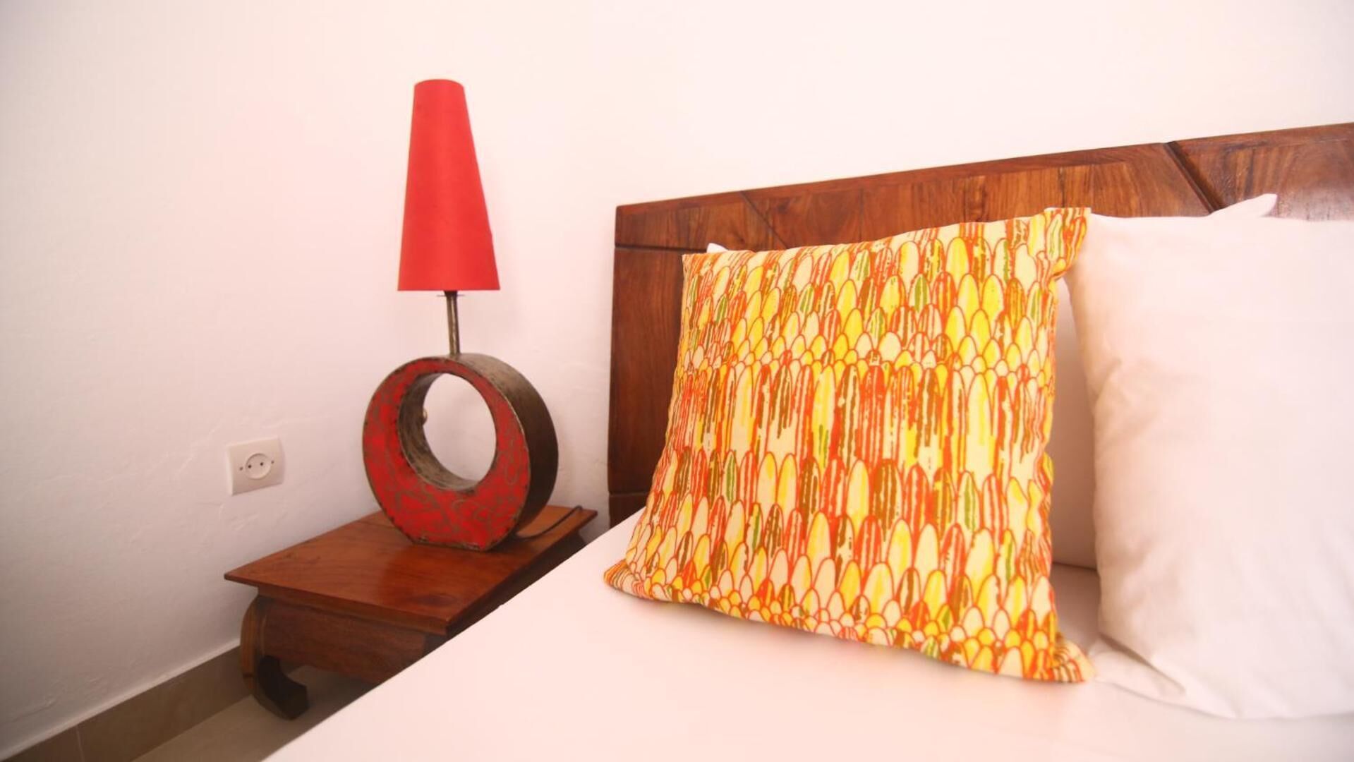 A bed with a patterned pillow, a plain pillow, and a red lampshade on a wooden nightstand.