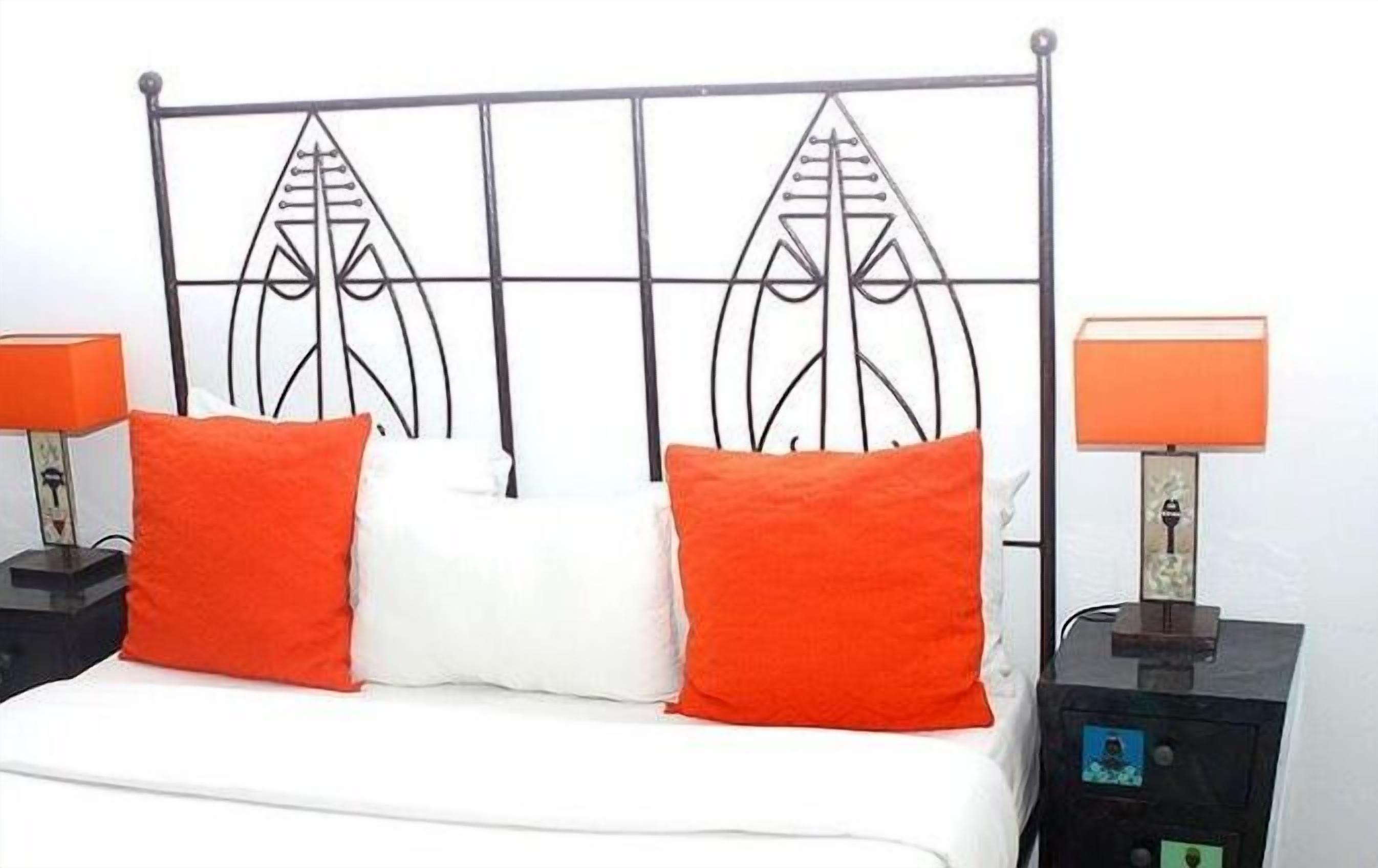 Ein Bett mit einem Metallkopfteil, zwei orangefarbenen Kissen und weißer Bettwäsche. Zwei Nachttische mit Lampen, auf einem befindet sich ein kleines elektronisches Gerät.