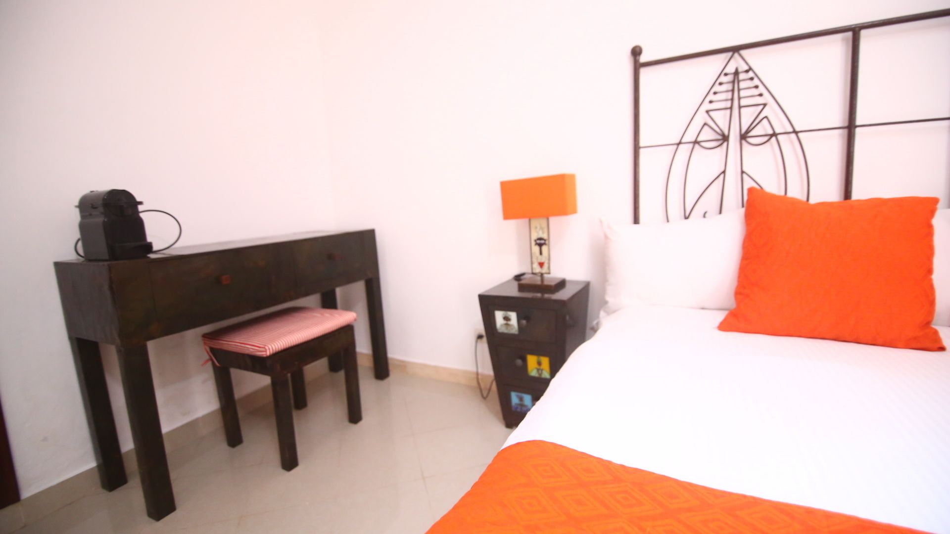 Un dormitorio con una cama, un cojín naranja, una mesita de noche con una lámpara, un banco de madera y un radiador fijado a la pared.