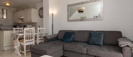 A modern living room with a grey sectional sofa, a white coffee table, and a dining area with white chairs and a small table.