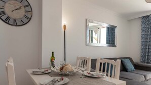 Un comedor con una mesa blanca dispuesta para dos, un reloj en la pared y un sofá gris al fondo.