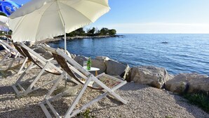 A beachside setting with wooden lounge chairs and a white umbrella, overlooking a calm sea and rocky shoreline. - Apartment Eva in Zambratija-Savudrija, air-conditioned, close to the beach (Umag/ Zambratija)