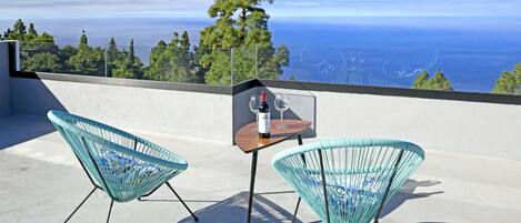 A balcony with two chairs and a small table, overlooking a scenic view of the sea and trees.