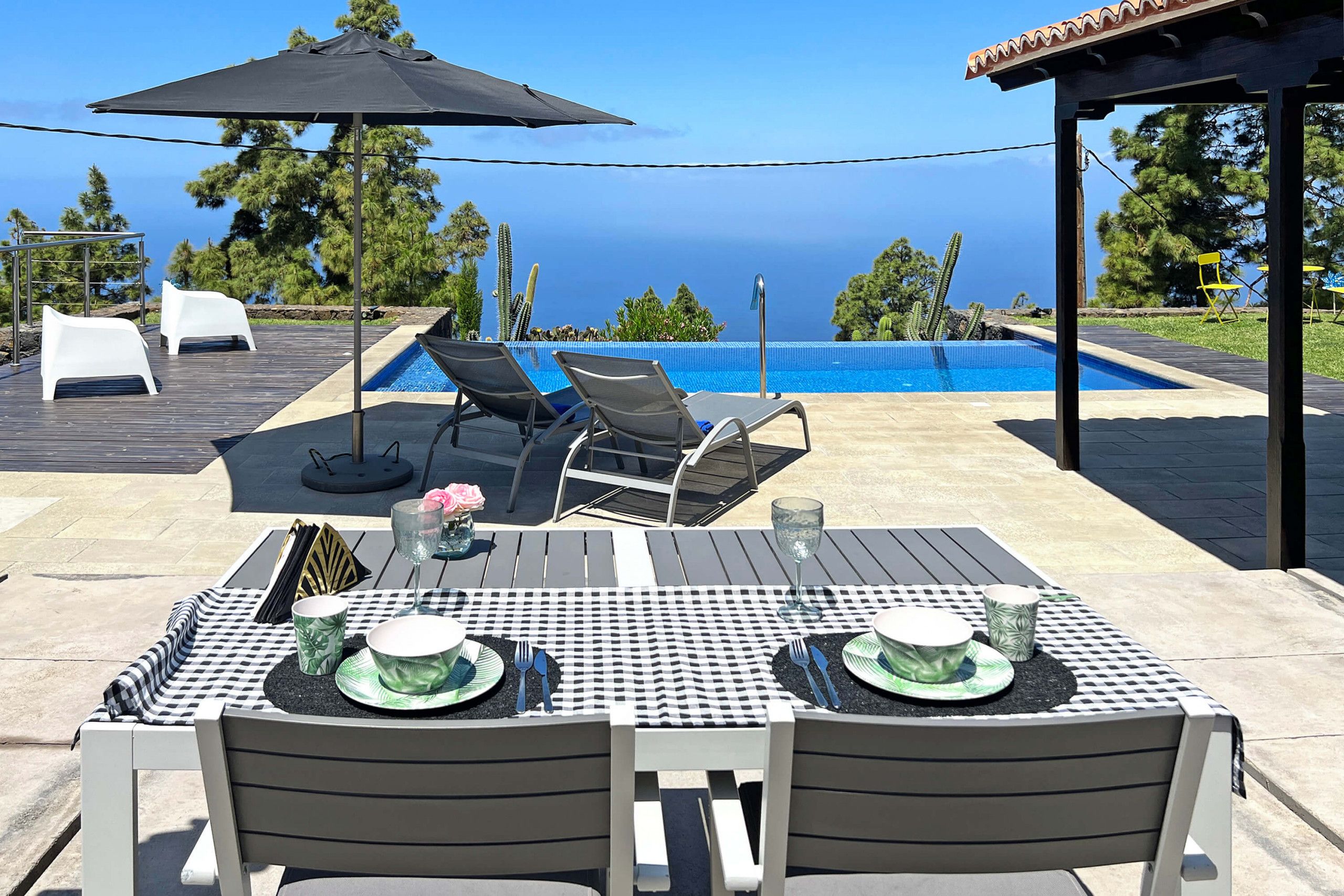 An outdoor pool area with lounge chairs, a table set for two, and a view of the ocean.