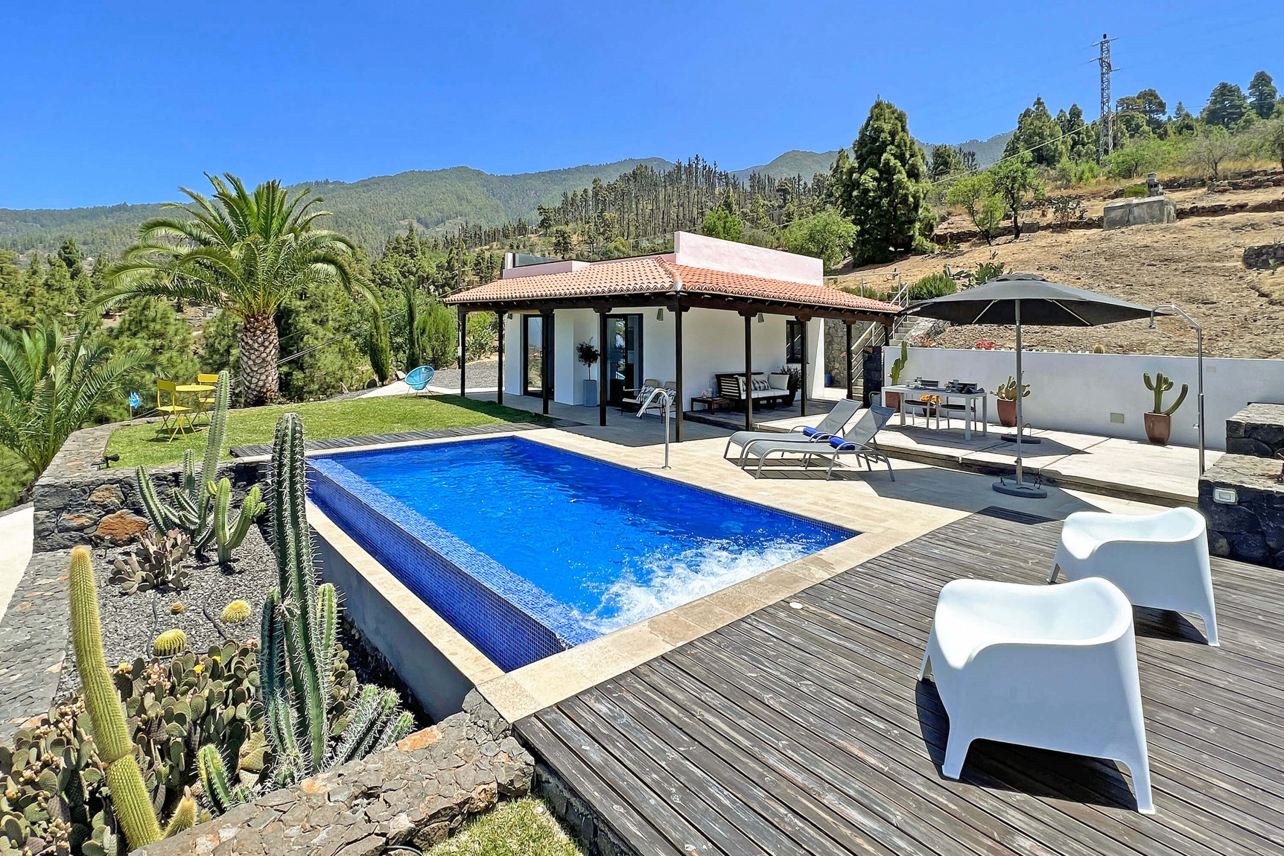 A modern outdoor pool area with a covered seating area, lounge chairs, and a view of a mountainous landscape.