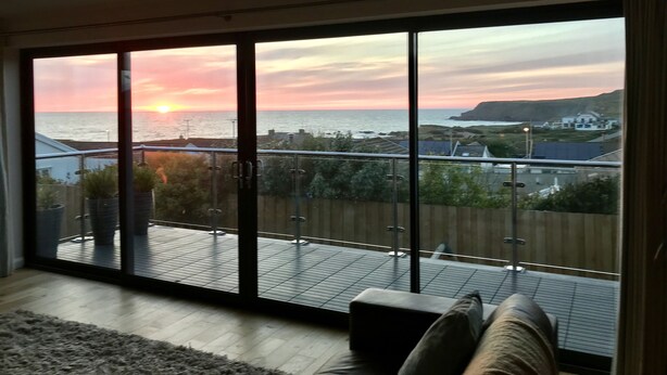 Een kamer met een grote schuifglazen deur die uitkomt op een balkon met uitzicht op zee en een zonsondergang.