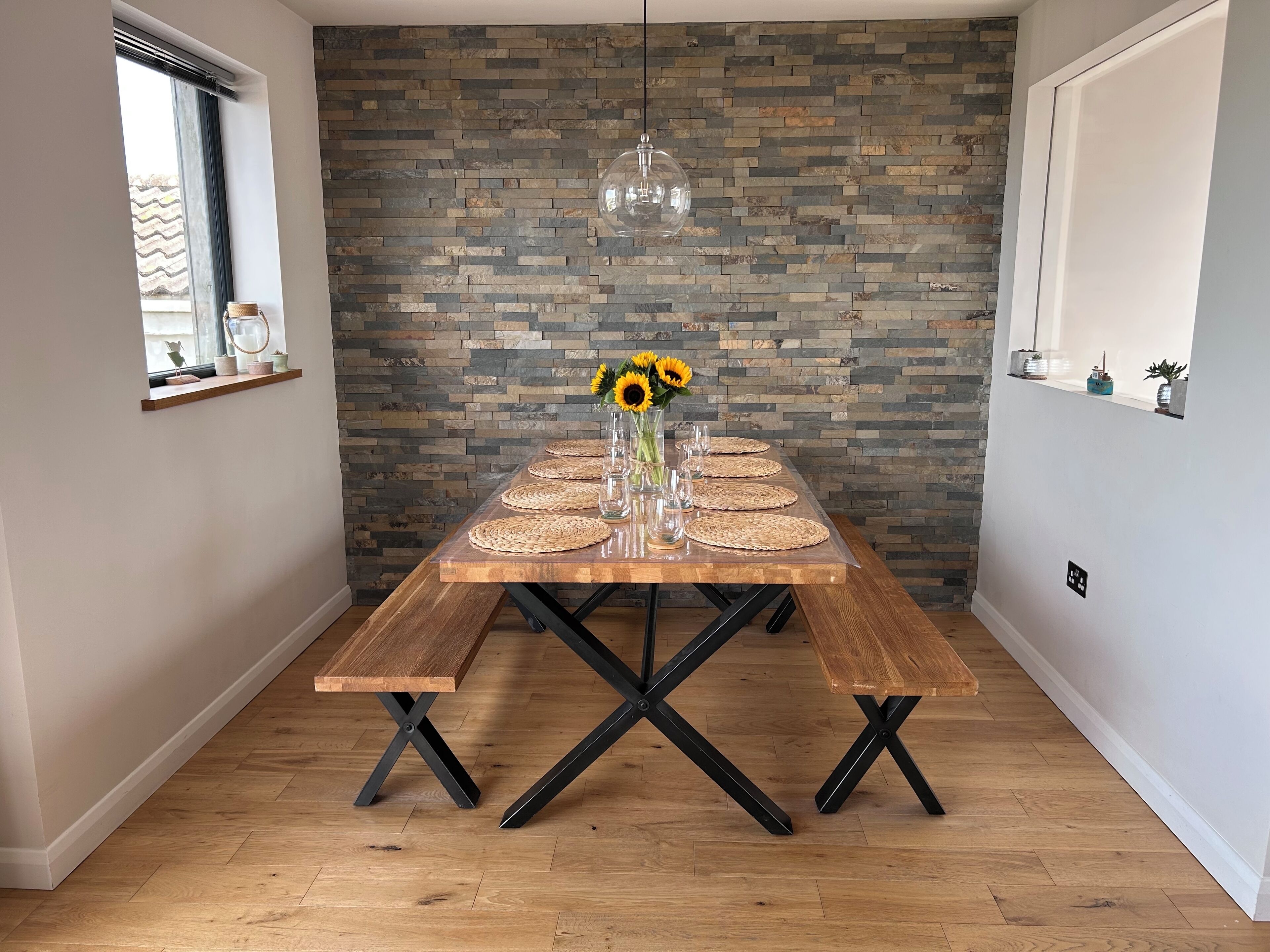 Un coin repas avec une table en bois, recouverte de sets de table en fibres tressées, de verres transparents et agrémentée de tournesols. Un mur de pierre avec un luminaire suspendu se trouve derrière la table.