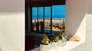 Een modern interieur met een groot raam met uitzicht op een strand. Op een witte plank staan drie potplanten.