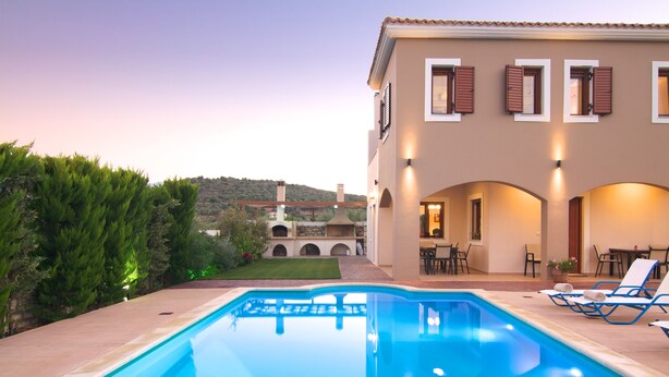 A swimming pool with lounge chairs, a patio area, and a building with arched windows and a tiled roof. - Villa Evanthia,a Place To Be! (Rethymno)