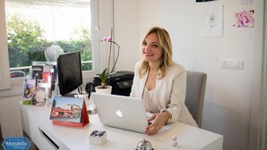 Uma mulher sentada à mesa com um laptop, rodeada por livros e uma pequena planta.
