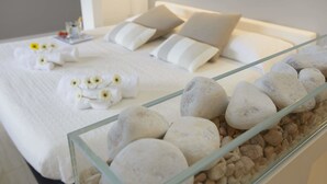 Cama com roupa de cama branca, almofadas e mesa de cabeceira de vidro com pedras decorativas.