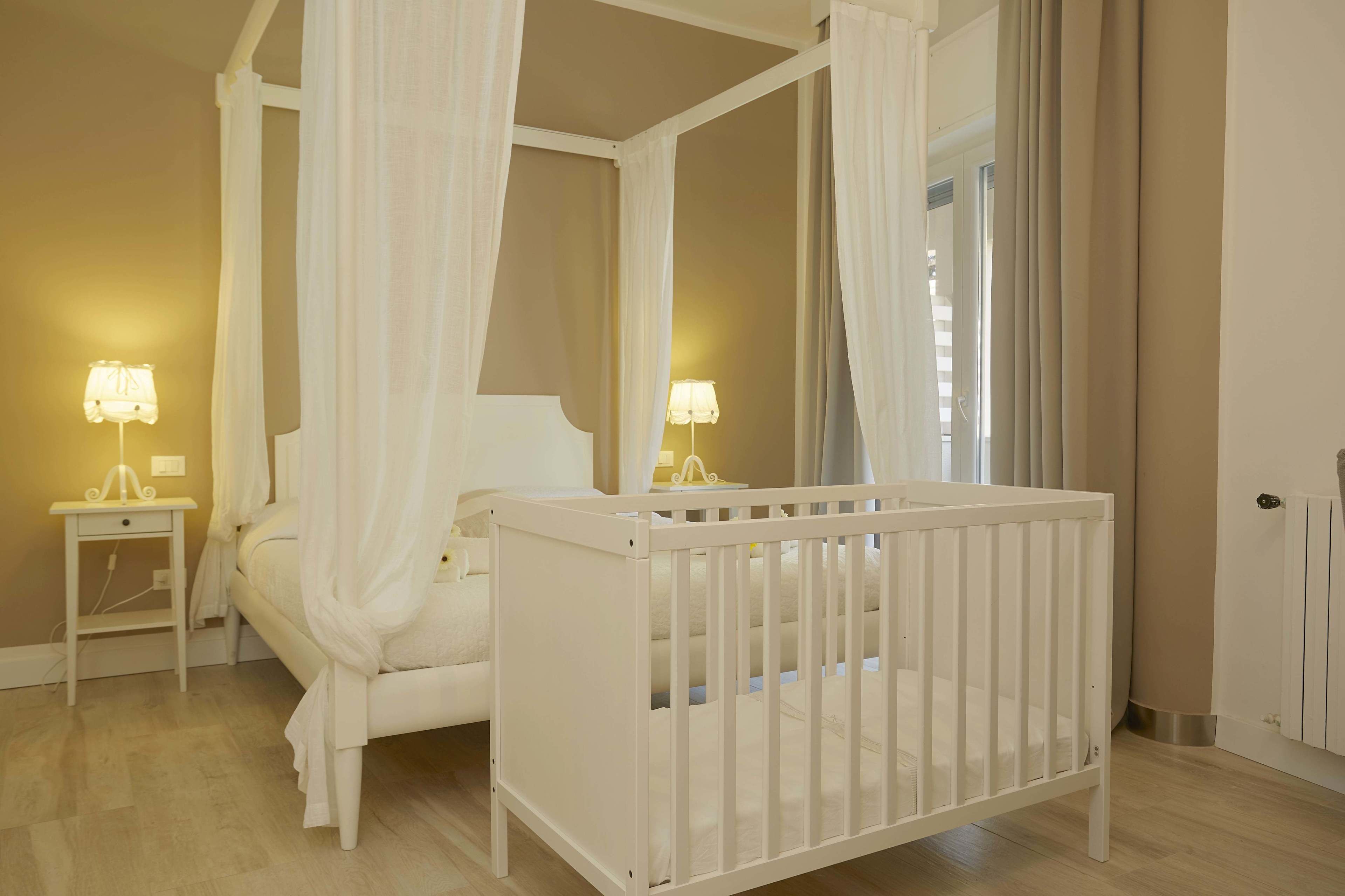 Una cama con dosel blanco, con cuatro postes, dos mesitas de noche con lámparas y una cuna blanca en una habitación con paredes color beige y suelo de madera.