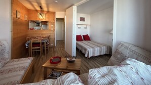 A cozy room with a wooden floor, a dining table with chairs, a bed with red pillows, and a small table with a basket and a book.