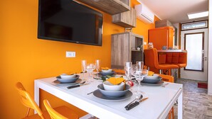A dining area with a white table set for four, orange chairs, a TV mounted on the wall, and a window with blinds.