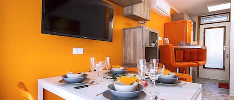 A dining area with a white table set for four, orange chairs, a TV mounted on the wall, and a window with blinds.