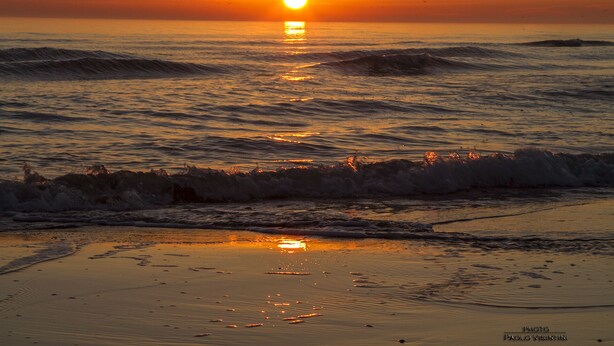 A beach at sunset with waves and a visible sun.