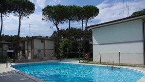 A swimming pool with a tiled deck, surrounded by trees and a building with a balcony.