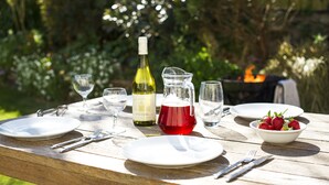 Una mesa de madera preparada para una comida, con platos, cubiertos, una botella de vino, una jarra de líquido rojo y un tazón de fresas.