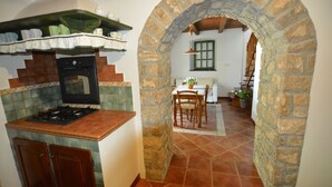 A kitchen with a stove, oven, and shelves with dishes. There is a dining area with a table and chairs, and a stone archway leading to a staircase.