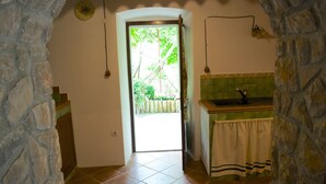 A narrow hallway with a stone wall on one side and a tiled floor. A door leads to a bright room with a view of greenery outside.