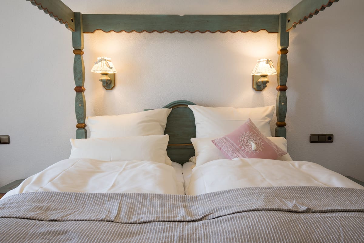 A neatly made bed with white linens and a striped blanket, a wooden headboard with a scalloped edge, and two wall-mounted lamps on either side.