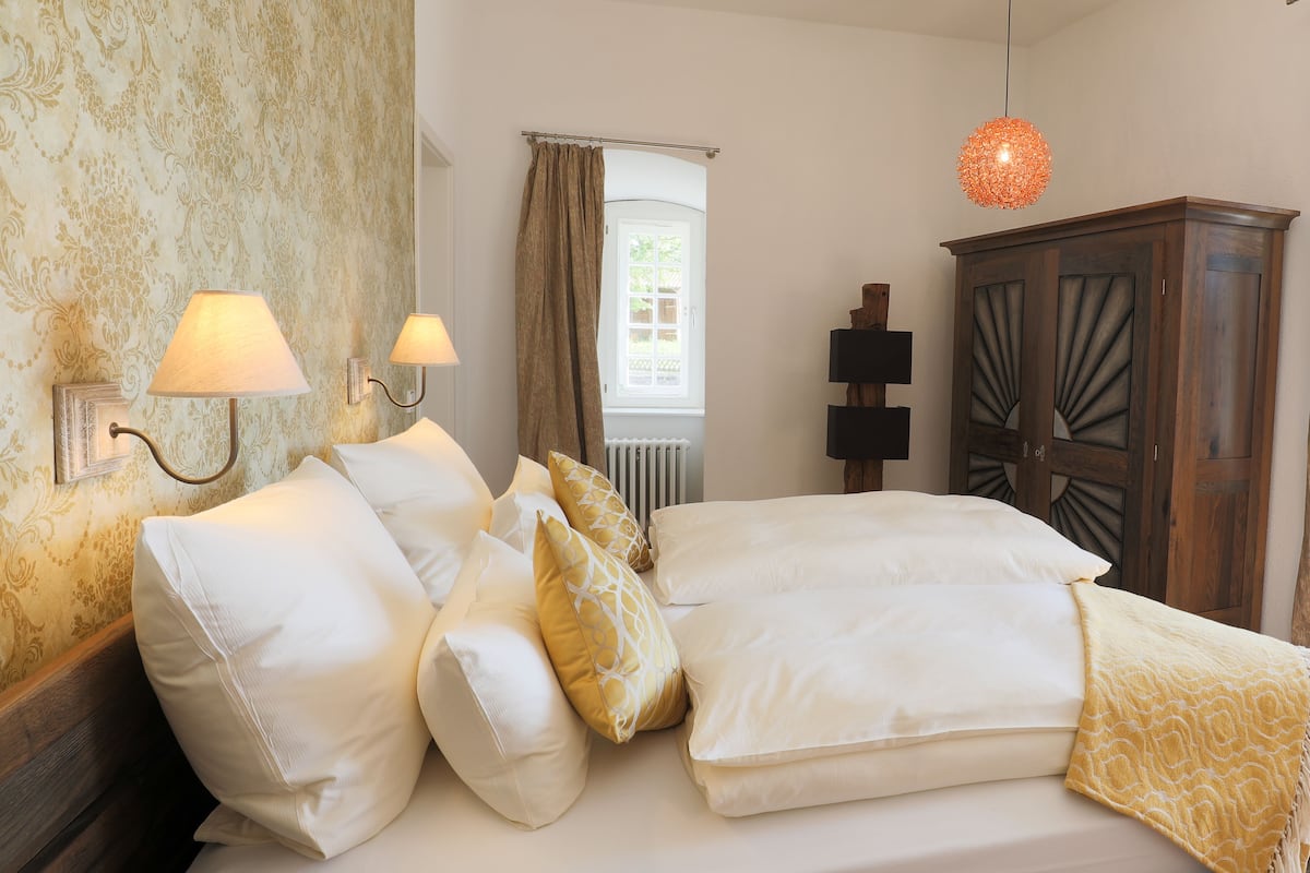 A bedroom with a bed, two wall-mounted lamps, a window with curtains, a radiator, and a wooden wardrobe.