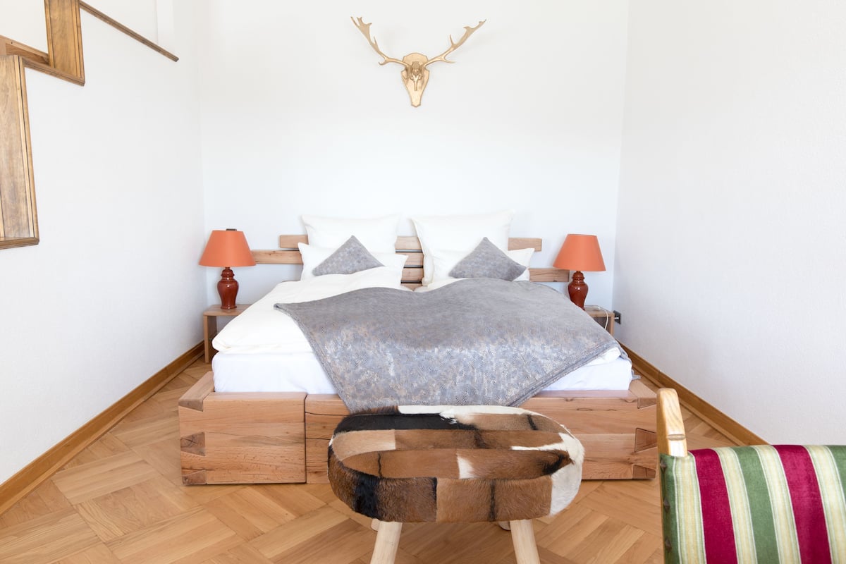 A bedroom with a bed, pillows, a wooden headboard, a wall-mounted deer skull, and a small wooden stool.