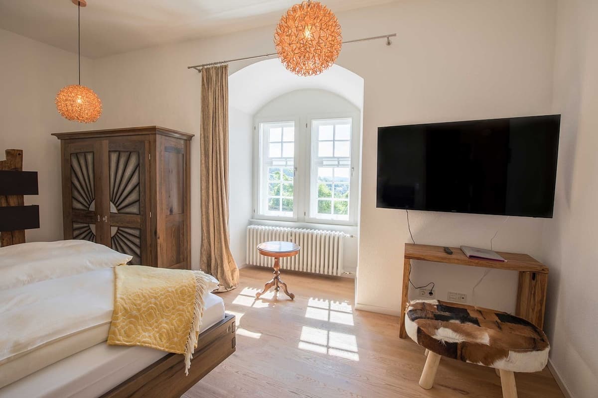 A bedroom with a bed, a wooden wardrobe, a television, a small round table, and a view of the outdoors through a window.