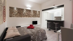 Una habitación de hotel moderna con una cama, un televisor, una obra de arte de piedra en la pared, una pequeña cocina y un baño visible a través de una puerta abierta.