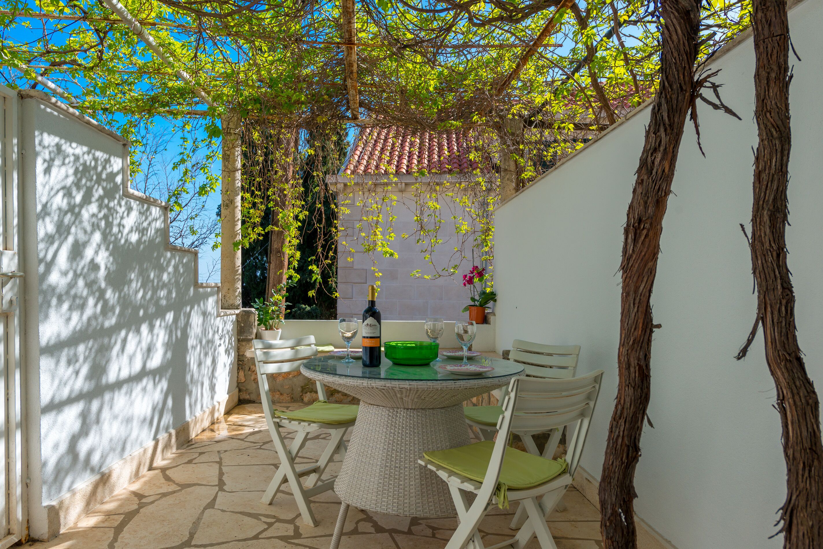 A round table with chairs, a bottle of wine, and a bowl in a covered outdoor area with white walls and a tiled floor.