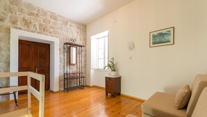 Un soggiorno con pavimento in legno, un divano beige, una porta in legno, una mensola metallica e una finestra con una pianta sul davanzale.