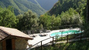 Un área de piscina con sillones de descanso, rodeada por un edificio de piedra con techo de tejas, frente a un telón de fondo de exuberantes montañas verdes.