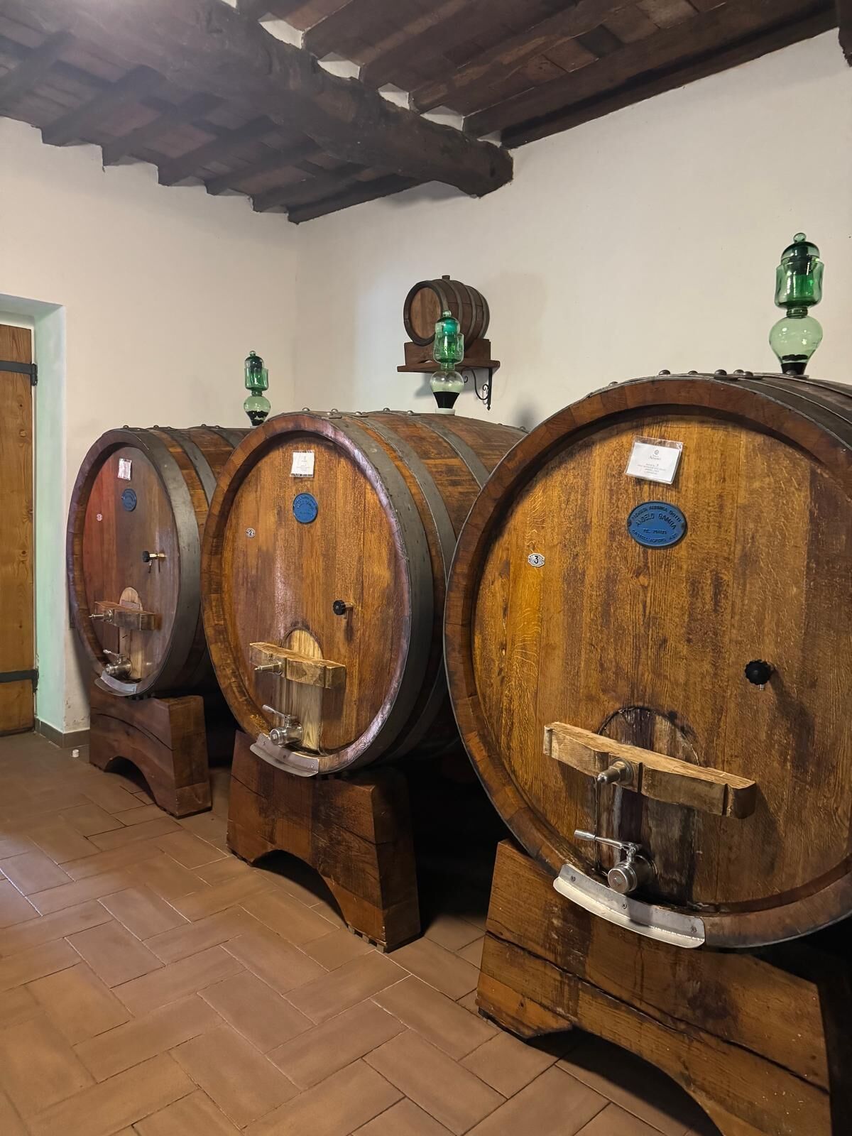 Three wooden wine barrels with metal spigots, arranged in a row in a room with wooden flooring and a tiled ceiling.