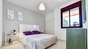 A bedroom with a bed, a nightstand, a lamp, a shelf with decorative items, and a window with a view of the outside. - Zafiro 18B - Front line apartment with sea views and direct access to the beach (Calpe / Calp)