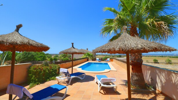 Un espace aménagé au bord de la piscine, avec des chaises longues et des parasols en chaume, un ciel d’un bleu éclatant et une vue imprenable sur le paysage.
