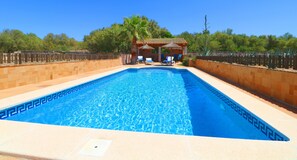 Une piscine rectangulaire bordée de carreaux, un palmier et un petit pavillon avec des chaises et une table.