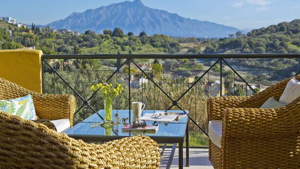 A balcony with wicker furniture, a table with a vase and a cup, and a mountain view in the background.