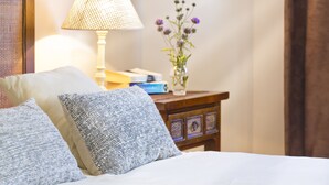 A neatly made bed with two pillows, a bedside table with a lamp, a vase with flowers, and a stack of books.