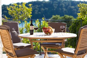 Een ronde houten tafel met twee stoelen, een fles wijn, een wijnglas en een schaal fruit op een balkon met uitzicht op bomen en heuvels.