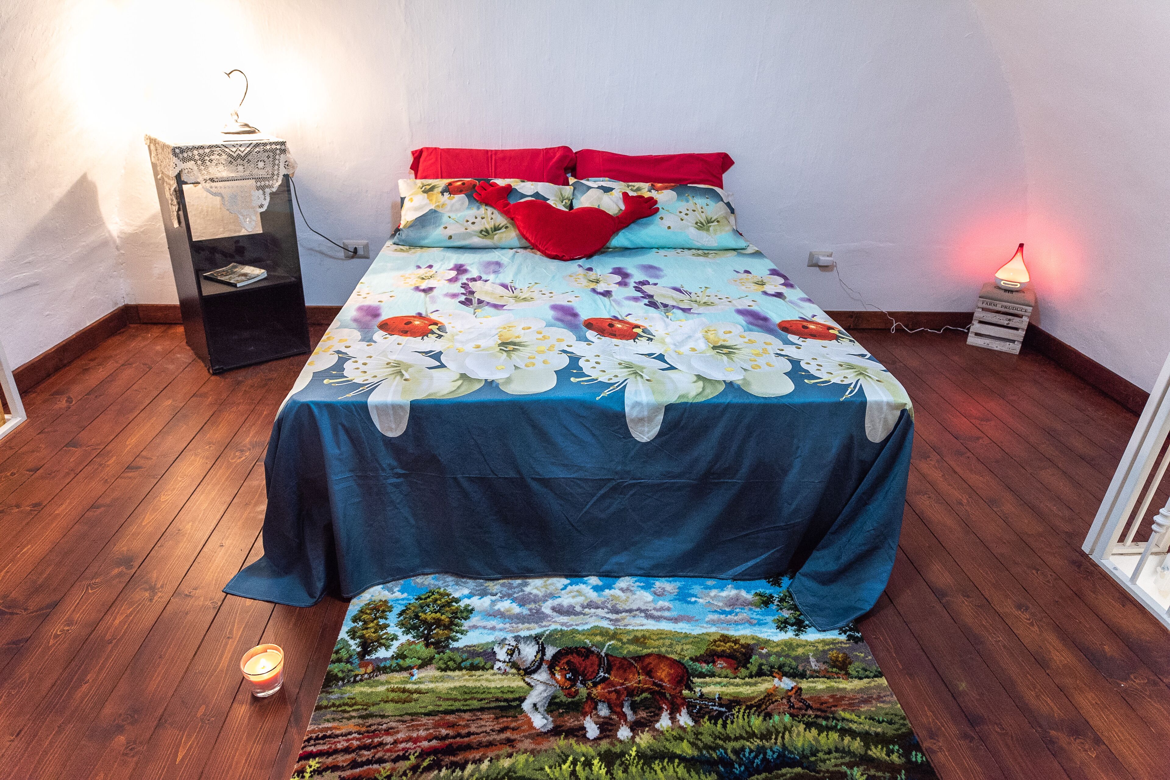 A bedroom with a floral bedspread, red pillows, a wooden floor, a bedside table with a lamp, and a rug with a horse scene.