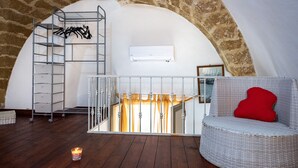 Un dormitorio tipo loft con techo alto, un equipo de aire acondicionado blanco, una silla de mimbre con cojín rojo, una escalera metálica y una vela encendida.