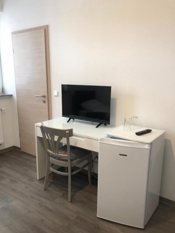 A small, neatly arranged room with a white desk, a chair, a television, and a compact refrigerator. - Hotel Vladi (Walldorf)
