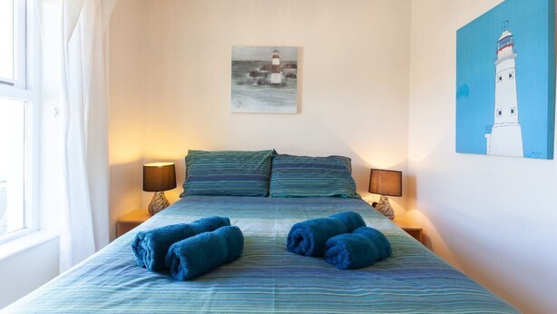 A bedroom with a bed, two bedside lamps, a painting of a lighthouse, and a window with curtains. - Portballintrae 10 Ballaghmore Cottages  (Bushmills)