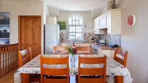 A compact kitchen with white cabinets, a tiled backsplash, and a wooden dining table with chairs. - Portballintrae 10 Ballaghmore Cottages  (Bushmills)