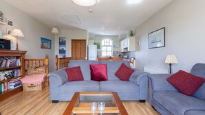 A spacious living room with a grey sofa, red cushions, a wooden coffee table, and a bookshelf filled with books. - Portballintrae 10 Ballaghmore Cottages  (Bushmills)