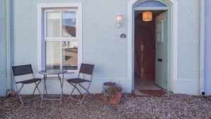 A building with a white exterior, a number '10' sign, a door with a light fixture, and two chairs outside. - Portballintrae 10 Ballaghmore Cottages  (Bushmills)